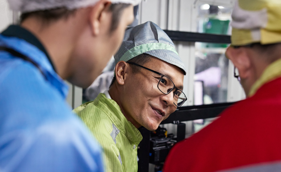 三名穿着工作服的员工在一台 AirPods Pro 组装机器旁交谈